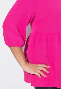 Top rosa con un tessuto strutturato, caratterizzato da maniche a sbuffo e una vita arricciata, mostrato su uno sfondo bianco semplice.