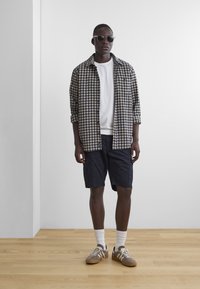 Casual outfit met een geblokt overhemd met knopen, een witte t-shirt, marineblauwe shorts en bruine sneakers met witte veters en geribde sokken.