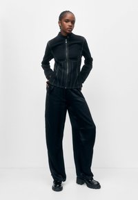 Veste noire zippée avec des coutures blanches contrastées, coupe ajustée et col montant. Portée avec un pantalon noir ample et des bottes noires épaisses.