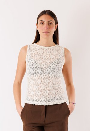 Donna con lunghi capelli scuri che indossa una canottiera bianca in pizzo e pantaloni marroni con le mani nelle tasche, in piedi contro uno sfondo semplice.