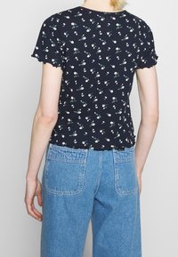 Top corto a maniche corte blu navy con orlo a scallop, decorato con motivi floreali bianchi e verdi, abbinato a jeans a vita alta azzurri.