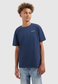 Tiener met krullend haar, gekleed in een marineblauwe "BALLIN'" t-shirt en lichtblauwe jeans, kijkt opzij tegen een effen achtergrond.