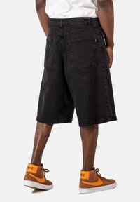 Pantalones cortos de mezclilla negra con un ajuste holgado, hasta la rodilla, dos bolsillos traseros y una cinturilla visible, combinados con zapatillas altas marrones.