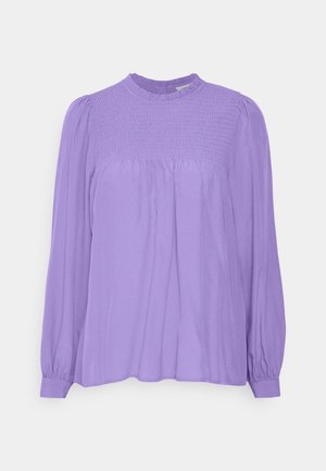 Blouse longue à manches longues en lavande avec empiècement smocké, col à volants, tissu froncé sous la poitrine et poignets boutonnés sur un fond uni.