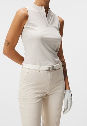 Kvinde iført ærmeløs beige top, beige bukser med hvidt bælte og en hvid golfhandske på højre hånd, mod ensfarvet baggrund.