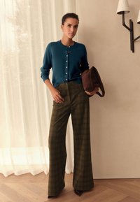 Tirkizno modra jopica z gumbi, kombinirana z olivno zelenimi širokimi hlačami z vzorcem kariranosti. Rjava torbica, elegantni čevlji z ostrim vrhom, nevtralno ozadje.