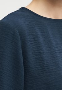 Εστιασμένη κοντινή εικόνα ενός ατόμου που φοράει μπλε-σκούρο τοπ από υφασμάτινο υλικό με στρογγυλεμένο λαιμό και ορατό λαιμό με λεία υφή.