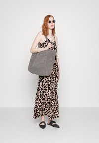 Geanta de pânză gri cu un buzunar, rochie maxi cu imprimeu leopard și pantofi slip-on negri. Modelul poartă ochelari de soare rotunzi. Fundal simplu.