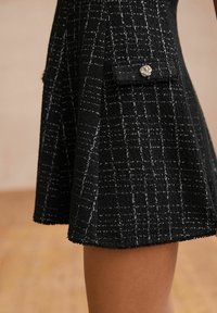 Jupe en tweed noire à motif quadrillé blanc, bouton torsadé doré sur la poche à rabat latérale, portée au-dessus du genou sur jambe nue.