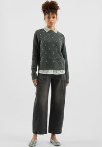Donna che indossa un maglione grigio con perline sopra una camicia di pizzo bianca, pantaloni neri a gamba larga e ballerine argentate glitterate, mentre si trova in piedi contro uno sfondo semplice.