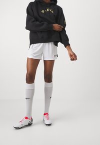 Sweat à capuche noir avec "PARIS" en jaune, short de sport blanc avec un logo Nike, chaussettes blanches jusqu'aux genoux et chaussures à crampons blanches avec des accents rouges.