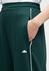 Ruka v vrecku tmavozelených tréningových nohavíc s bielym pruhom a elastickým pásom, zobrazujúca malú bielu logo na stehne.