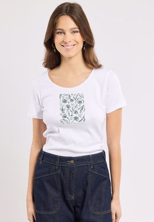 Femme souriante, portant un T-shirt blanc avec un motif floral vert et un jean taille haute bleu foncé, debout devant un fond clair uni.