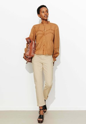 Femme aux cheveux courts portant un chemisier marron et un pantalon beige, tenant une veste à motifs, debout devant un fond blanc, chaussée de sandales noires.