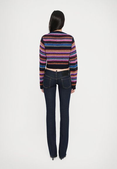 Suéter corto de rayas multicolores con hilos brillantes, combinado con jeans de campana en azul oscuro que presentan una etiqueta en la cintura y líneas limpias.