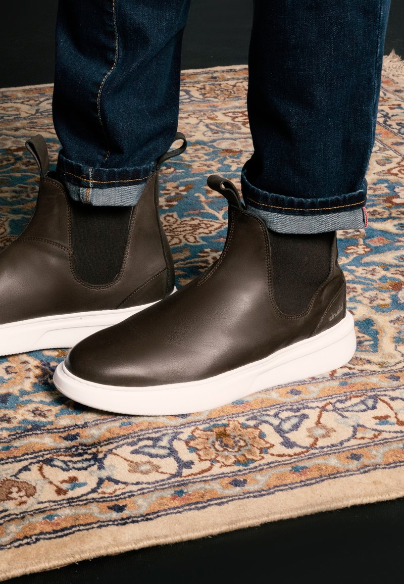 Botas de cuero marrón sin cordones con paneles elásticos en los laterales, suelas de goma blancas y detalles de costura sutiles, combinadas con unos jeans oscuros doblados.