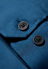 Blauwe stof met een gestructureerde weving, met twee zwarte knopen op de kraag. Genaaide randen zijn zichtbaar, wat de ambachtelijke details benadrukt.