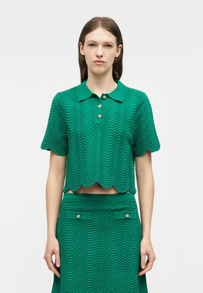 Γυναίκα που φοράει πράσινη υφασμάτινη κοντομάνικη πόλο μπλούζα με κυματιστές άκρες και ταιριαστή φούστα με χρυσά κουμπιά μπροστά.