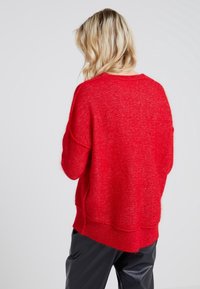 Roter gestrickter Pullover mit lässiger Passform, rundem Halsausschnitt und geripptem Saum. Weiche Textur mit lockeren Ärmeln, getragen über schwarzen Hosen.