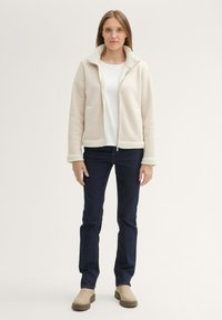 Veste zippée beige clair avec un col et des poignets doux et pelucheux, associée à un jean bleu foncé et des bottines beiges. Deux poches avant visibles.