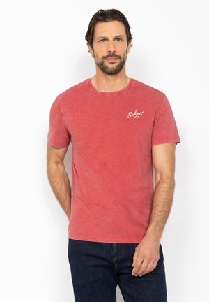 Mand med brunt hår og skæg iført falmet rød kortærmet t-shirt og mørkeblå jeans, står mod hvid baggrund.