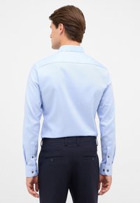 Camicia azzurra chiara con maniche lunghe, polsini bottonati e vestibilità su misura, abbinata a pantaloni navy scuri. Tessuto liscio, design classico.