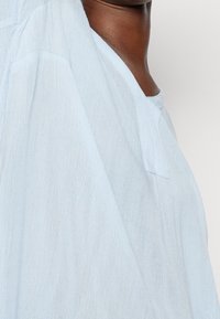 Nahaufnahme einer Person, die ein lockeres, hellblaues, strukturiertes Kleidungsstück mit weit ausgeschnittenem Halsausschnitt vor einem schlichten Hintergrund trägt.