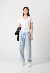 Vit V-ringad t-shirt, lösa ljusblå denimjeans och vita sneakers. Svart sweatshirt med text som hålls i ena handen. Enkelt, avslappnat outfit.
