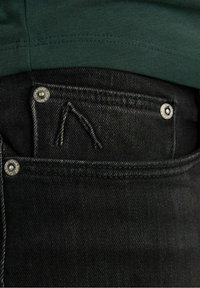 Černé džíny z denimu s přední kapsou a výrazným prošíváním a kovovými knoflíky. Tmavě zelená košile je částečně viditelná nad džíny.