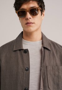 Brown herringbone button-up jacket over a light gray ribbed sweater. Dark sunglasses with a tan frame. Plain background.