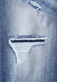 Niebieska tkanina dżinsowa z trójkątnym rozdarciem z postrzępionymi krawędziami, odsłaniającym ciemniejszą, niebieską warstwę pod spodem; tekstura jest miękka i zużyta.