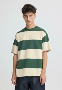 T-shirt à manches courtes rayé vert et beige, fabriqué en tissu doux avec une coupe décontractée et un col rond, porté avec un jean foncé.