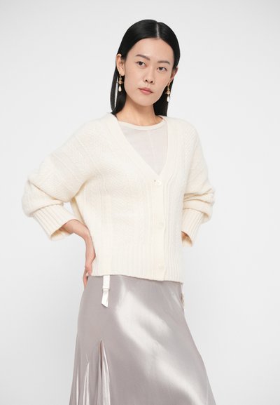 Krémový kardigan s texturou, výstřihem do V, zapínáním na knoflíky a širokými rukávy, kombinovaný s lesklou šedou sukní se bočními rozparky.