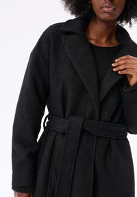 Czarny płaszcz z mieszanki wełny z fakturowaną powierzchnią, szerokimi klapami i zawiązywanym paskiem w talii, pokazany od pasa w górę na modelce.