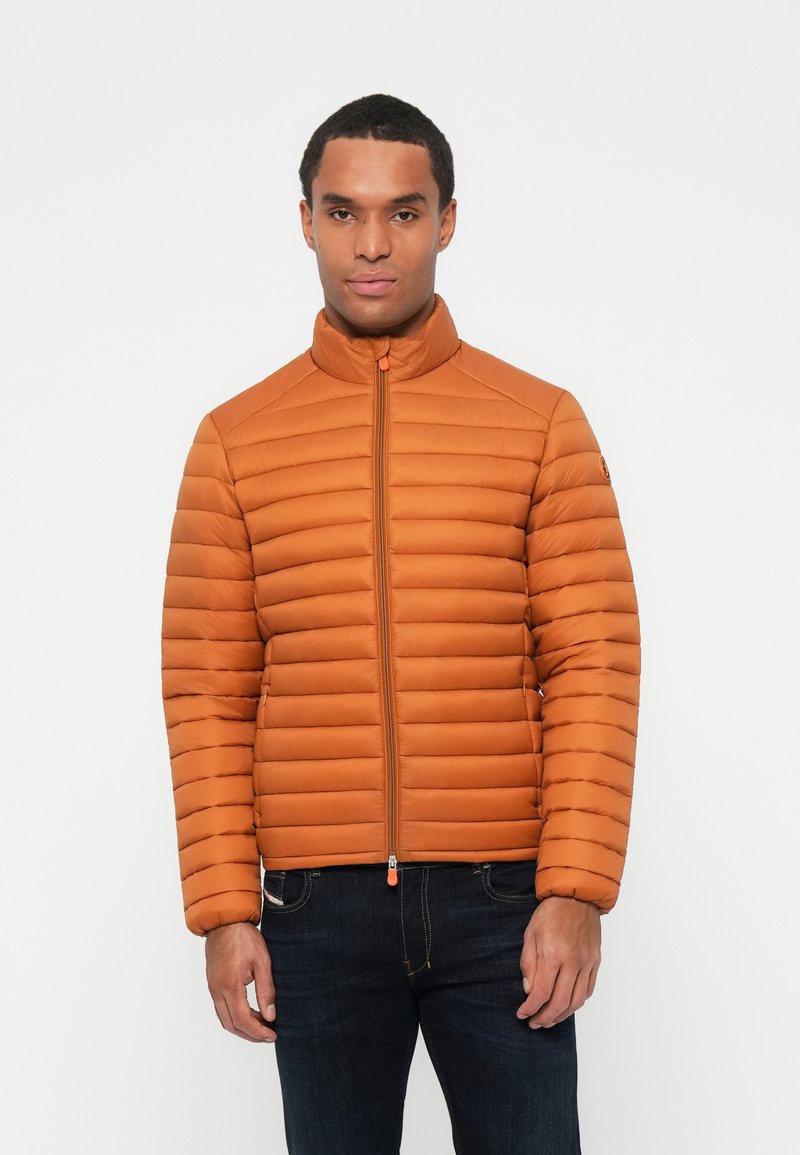 Veste rembourrée orange avec un col montant, un matelassage vertical et une fermeture éclair à l'avant. Associée à un jean bleu foncé pour un contraste.