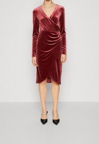 Femme portant une robe cache-coeur en velours rose poussiéreux, à manches longues et longueur genoux, ornée de détails froncés, associée à des escarpins noirs à bout pointu avec des brides en perles.