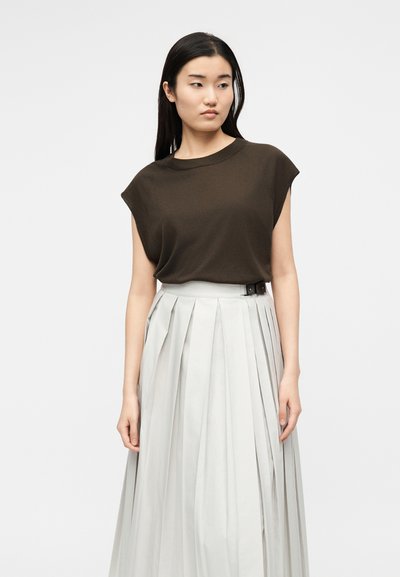 Femme aux longs cheveux noirs portant un haut sans manches marron foncé et une jupe plissée taille haute gris clair, debout sur fond blanc.