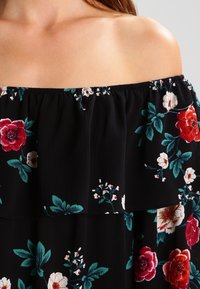 Svart off-shoulder topp med blommönster i rött, vitt och grönt, med en volangkrage och textilier med struktur.
