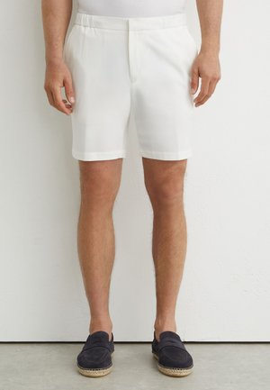 Uomo con pantaloncini bianchi su misura e espadrillas nere slip-on, in piedi contro un muro bianco e un pavimento grigio.