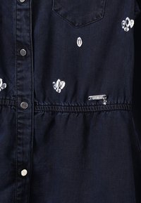 Mustat farkkukangasvaate, jossa on hopeiset nepparit, kirkkaat koristetähdet, rintatasku ja pieni metallinen "Guess"-brändilogo.