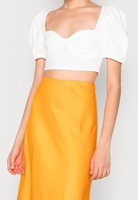 Bílý zkrácený top s nafouknutými rukávy a výstřihem do srdce, doplněný jasně žlutou sukní s vysokým pasem s hladkou texturou.