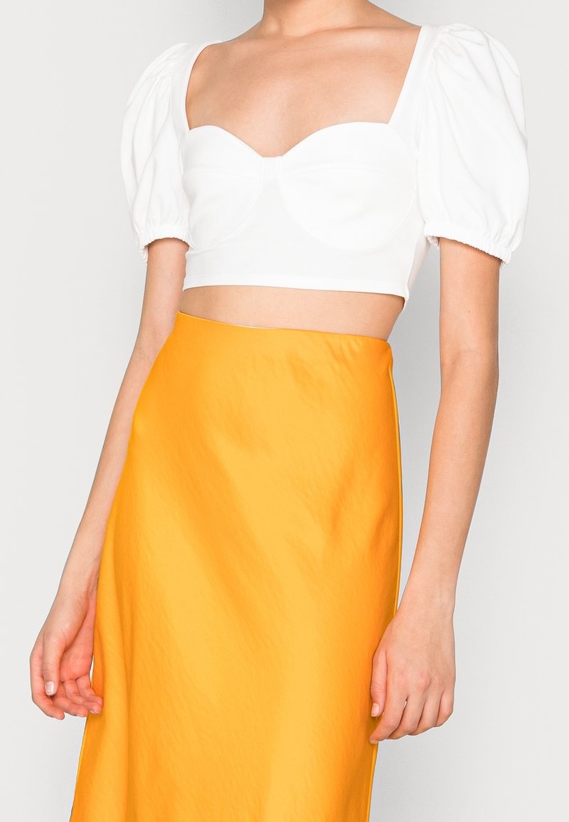 Bílý zkrácený top s nafouknutými rukávy a výstřihem do srdce, doplněný jasně žlutou sukní s vysokým pasem s hladkou texturou.