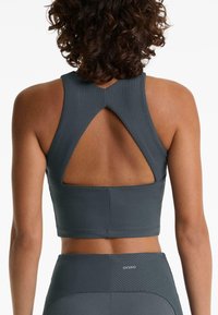 Femme aux cheveux bouclés portant un soutien-gorge de sport texturé gris foncé avec une découpe triangulaire dans le dos et un legging taille haute assorti.