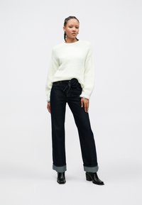 Hvit strikkegenser, mørkeblå vidbukser med blomsterformet beltedetalj, og svarte ankelstøvler med hæl. Jevn tekstur, avslappet passform.