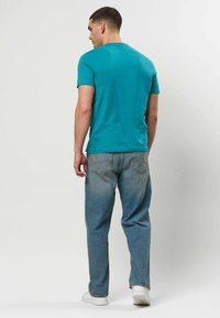 Homme portant un t-shirt turquoise à manches courtes, un jean bleu ample et des baskets blanches, debout de dos devant un fond clair uni.