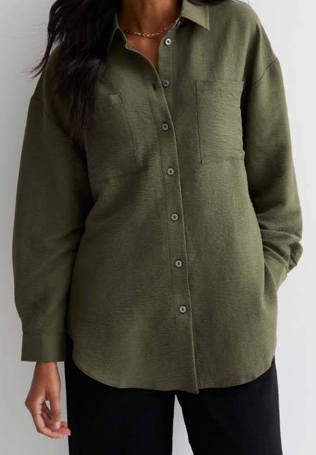 Kvinna i olivgrön knappar-skjorta med två bröstfickor och svarta byxor, stående med ena handen i fickan.
