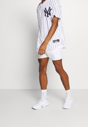 Persona vestida con zapatillas deportivas blancas, pantalones cortos blancos y una camiseta de béisbol de los New York Yankees con rayas blancas, de pie frente a un fondo liso.