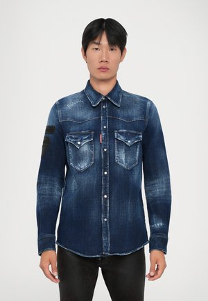 Giovane in piedi, che indossa una camicia di jeans blu scuro scolorita con tasche a patta con bottoni e pantaloni neri, su uno sfondo semplice.