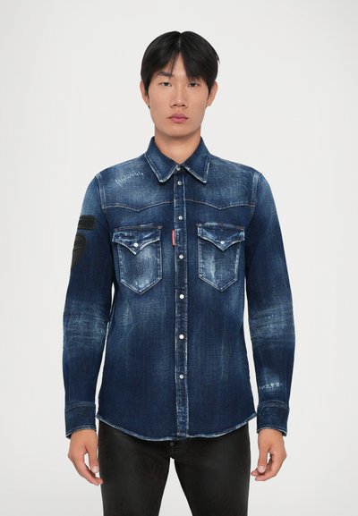 Young man standing, wearing a dark blue faded denim shirt with buttoned flap pockets and black pants against a plain background.