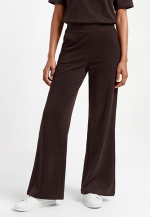 Kobieta ubrana w luźne, szerokie spodnie w ciemnym brązowym kolorze oraz białe trampki, stojąca na tle jednolitego jasnego tła.